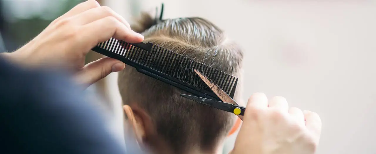 A close up picture of a young man getting his hair cut. Currently, the boy's brown hair is being filtered through a comb, while scissors cut the hair that's in the comb, giving a nice, close cut.