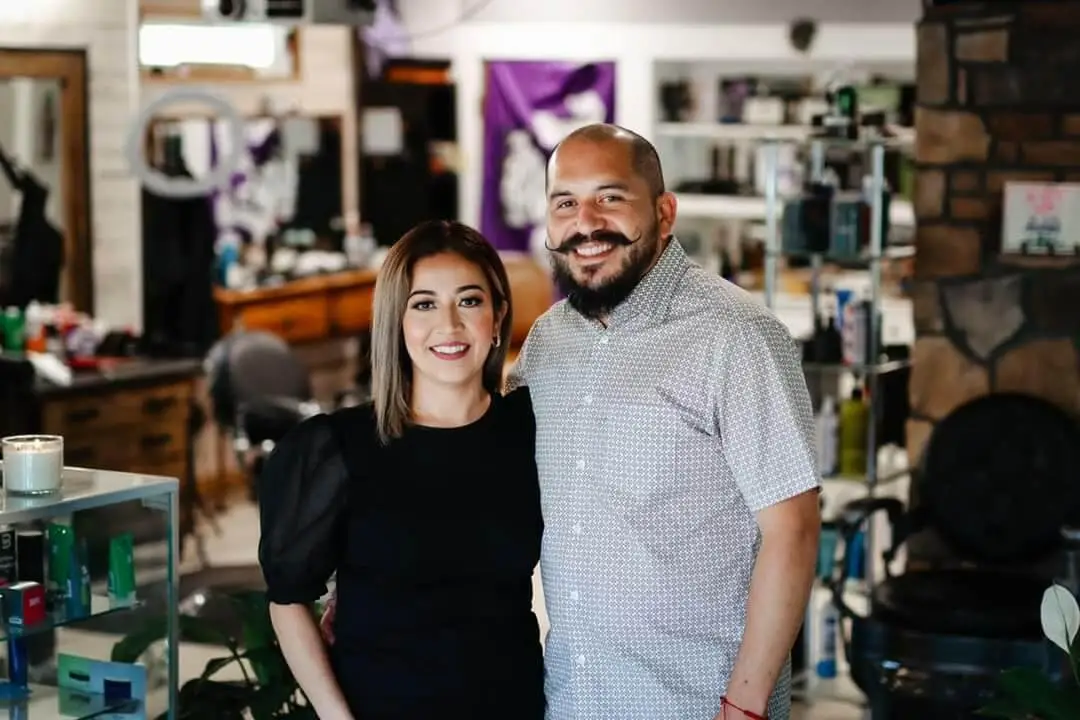 An image of Rudy the barber with his wife at his side. Rudy is tall, with a clean, shaven head, and grown out facial hair, with  and his mustache is  is curled at the end. His wife is about a foot smaller than him, her hair is in a short bob, and she is wearing a black blouse. They are both in the foreground, in the background is the rest of the barber shop, but blured.